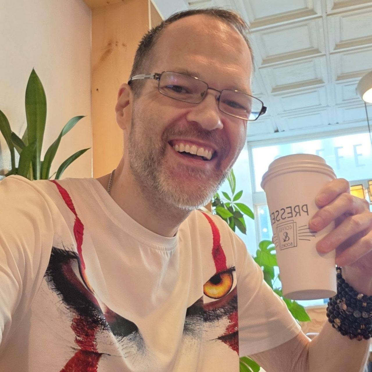 David Strausser in a white tiger-print t-shirt holding a Pressed Coffee and Books cup in a bright cafe