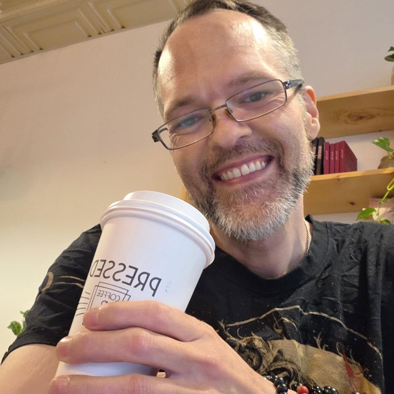 David Strausser holding a Pressed Coffee and Books cup in a black t-shirt, wooden bookshelves behind him